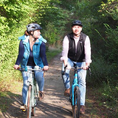 Bild vergrößern: Ein Mann und eine Frau fahren mit ihren Fahrrdern einen Waldweg entlang. Die Frau blickt den Mann lchelnd an. Beide tragen Fahrradhelme.