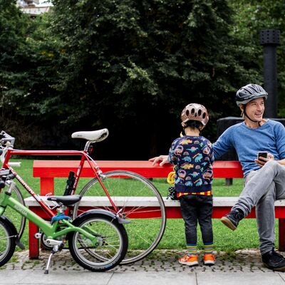 Bild vergrößern: Ein Mann mit Fahrradhelm sitzt auf einer roten Bank, der Kamera zugewandt neben ihm steht ein Kind, welches ebenfalls einen Fahrradhelm trgt. An die Bank sind ein rotes Herrenrad und ein grndes Kinderrad gelehnt. Im Hintergrund ist eine Wiese und ein Wald zu sehen.