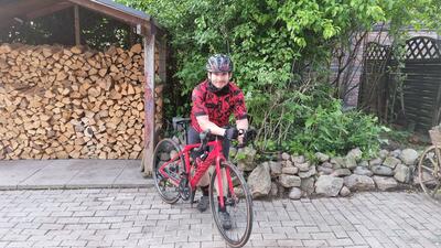 Bild vergrößern: Ein junger Mann mit Fahrradhelm steht neben einem roten Fahrrad und sttzt sich mit beiden Unterarmen auf dem Lenker auf. Er blickt lchelnd in die Kamera. Im Hintergrund ist gestapeltes Brennholz und Bepflanzung zu sehen.