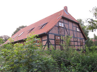 Bild vergrößern: Auf dem Bild ist ein Fachwerkhaus mit roten Backsteinen und rotem Satteldach zu sehen, welches von Bumen und Struchern umgeben ist.