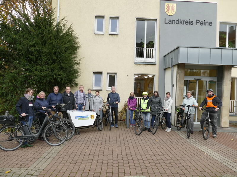 Mehrere Frauen und Männer stehen vor einem Gebäude, an dem das Wappen und der Schriftzug des Landkreises Peine zu sehen sind und blicken in die Kamera. Einige der Personen stehen neben Fahrrädern.