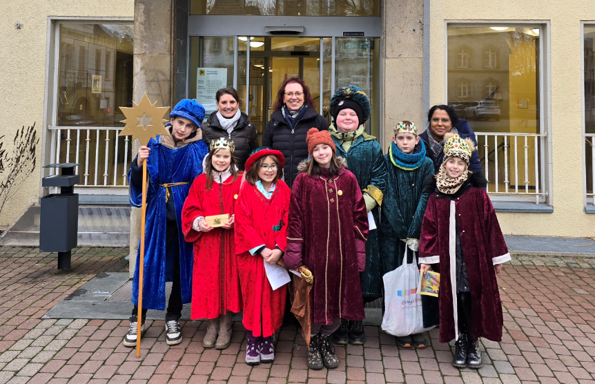 Drei Frauen und eine Gruppe von Mädchen und Jungen stehen vor einem Gebäude und lächeln in die Kamera. Die Mädchen und Jungen sind als Sternsinger gekleidet.