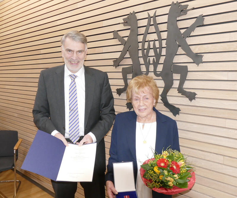 Ein Mann und eine Frau stehen vor einer vertäfelten Wand, die das Kreiswappen in schwarz zeigt. Der Mann hält eine Urkunde in den Händen und die Frau einen Blumenstrauß.